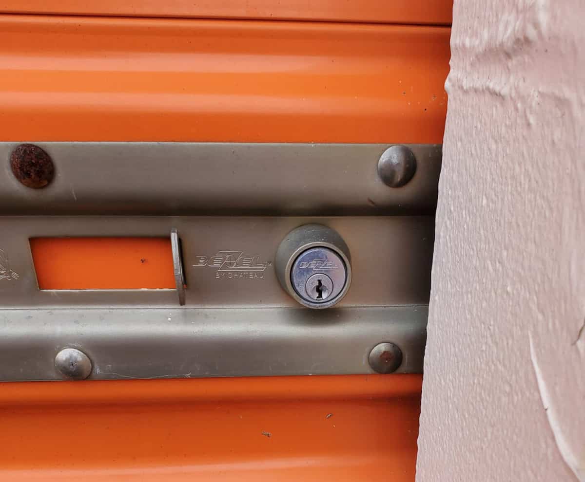 Close-up of a metal latch with a cylinder lock and key on an orange storage unit door next to a beige wall in New Jersey.