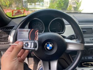 A hand holds a BMW key fob and a business card from a locksmith near me in front of a BMW steering wheel and dashboard.