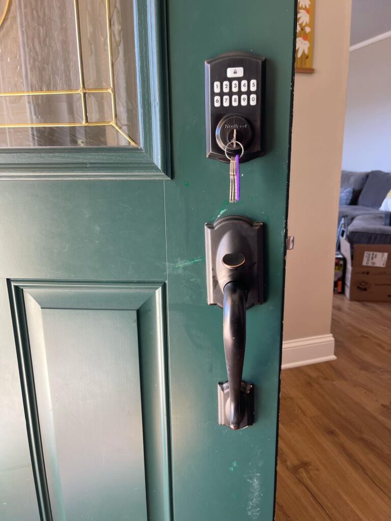 Green front door slightly open, showing a keypad lock with a key hanging from it and a black handle, with part of a living room visible inside—perfect for New Jersey homes seeking secure entry solutions.