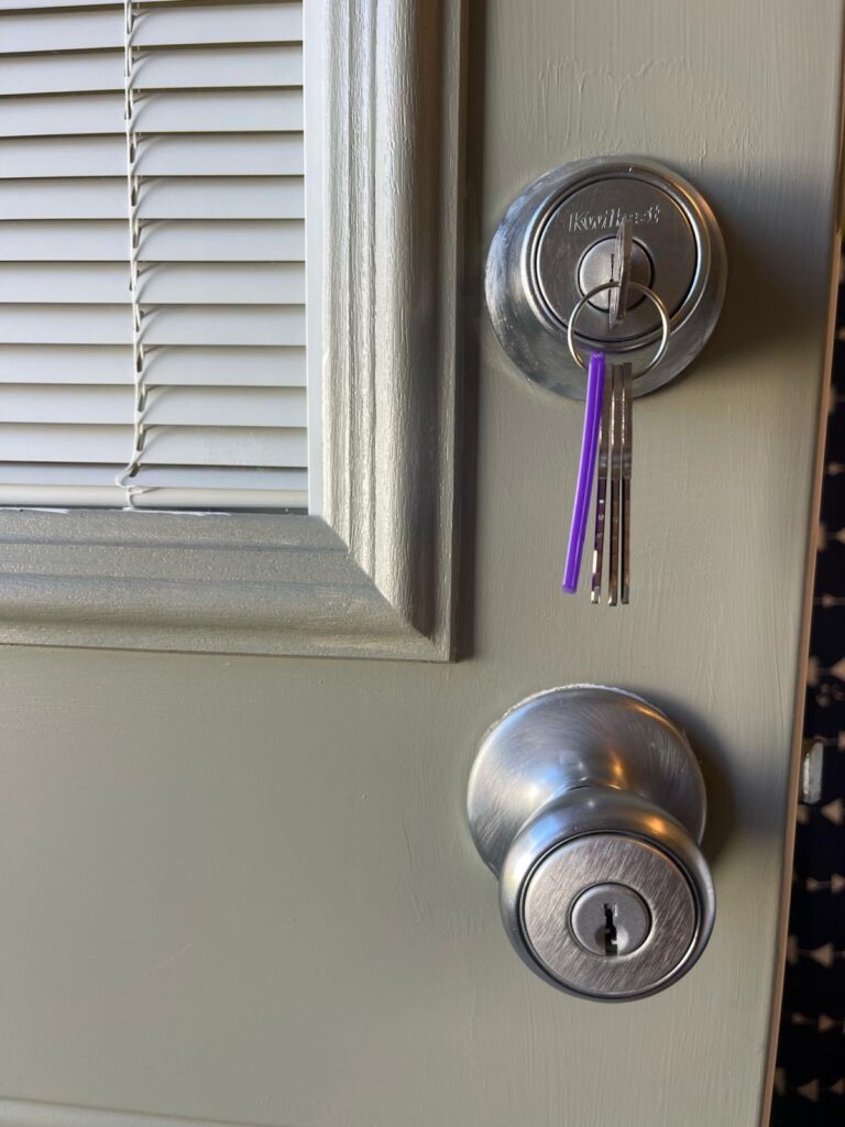A set of keys with a purple keychain is inserted in a deadbolt lock on a closed beige door, perfect for anyone searching "locksmith near me," with blinds covering a nearby window.