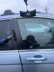 A person uses inflatable pump wedges to open the door of a blue car with a key on a rainy day in New Jersey. The window is wet with raindrops and the person's reflection is visible in the glass.