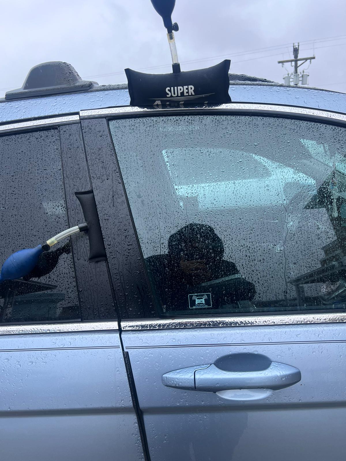 A person uses inflatable pump wedges to open the door of a blue car with a key on a rainy day in New Jersey. The window is wet with raindrops and the person's reflection is visible in the glass.