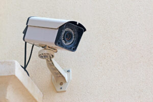 A security camera, expertly installed by a New Jersey locksmith, is mounted on a beige exterior wall, angled downward, with visible wires and a sturdy mounting bracket resembling a large key.