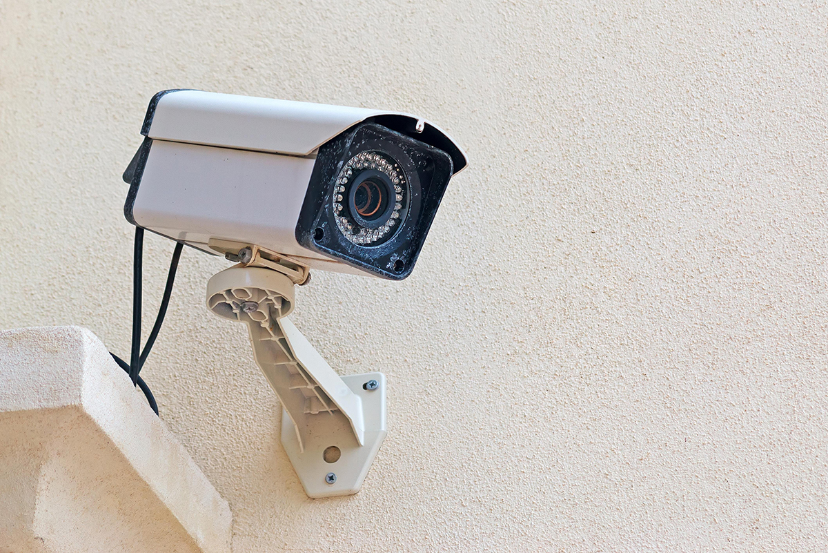 A security camera, expertly installed by a New Jersey locksmith, is mounted on a beige exterior wall, angled downward, with visible wires and a sturdy mounting bracket resembling a large key.