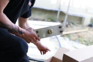 A person kneels outdoors beside an open cardboard box in New Jersey, holding a rectangular metal plate with bolts and a central fastener—tools often used by a locksmith as part of their key-fitting work.