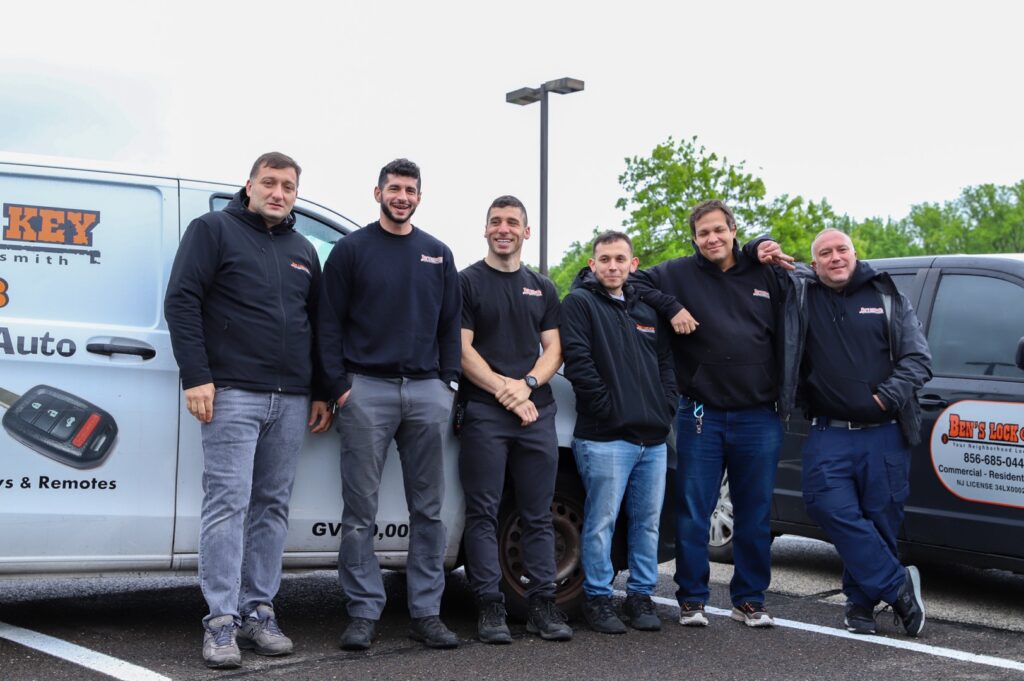 Six locksmiths in black uniforms stand beside two company vans in a New Jersey parking lot, posing for a group photo on a cloudy day, each proudly holding a key.