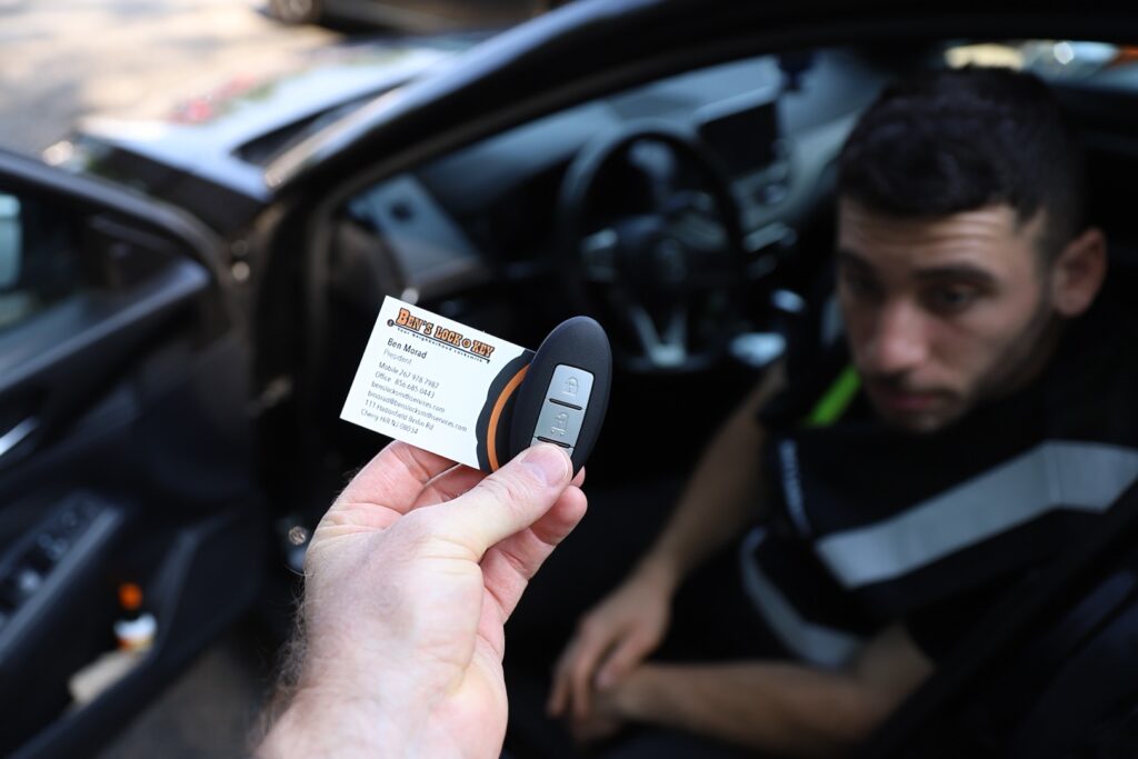 A hand holding a key fob and a business card in front of a local man sitting in the driver’s seat of a car with the door open.