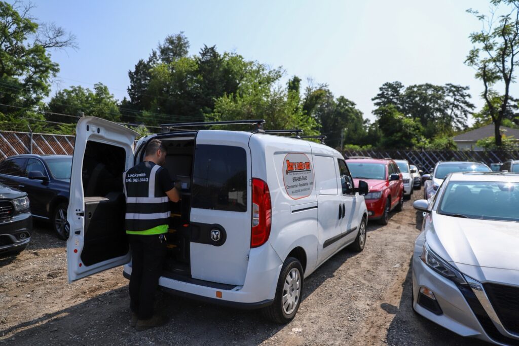 A person stands at the open back doors of a white locksmith van parked in a lot surrounded by other vehicles on a sunny day in New Jersey.