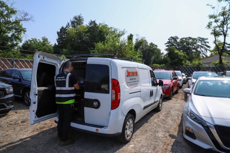 A person stands at the open back doors of a white locksmith van parked in a lot surrounded by other vehicles on a sunny day in New Jersey.