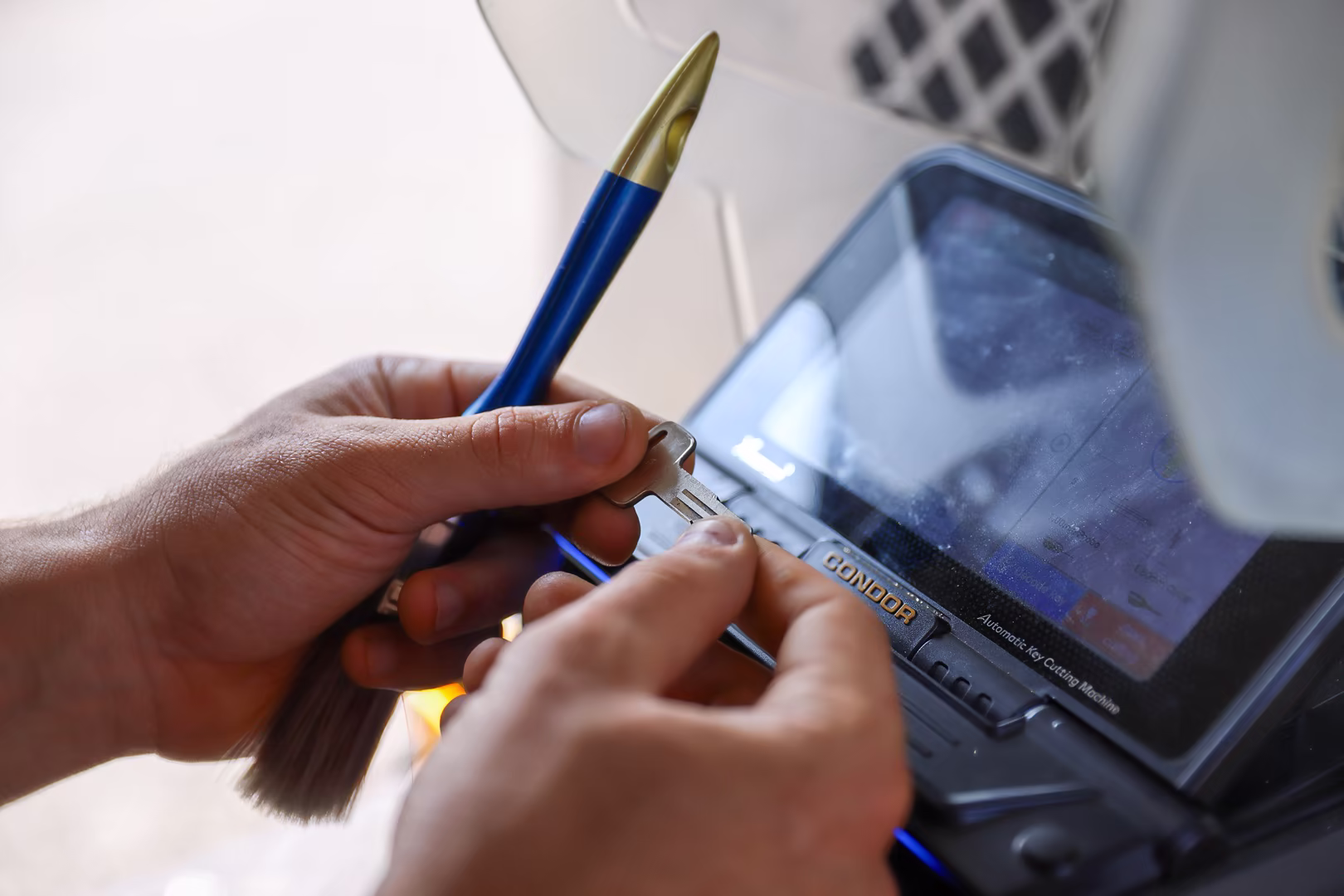 A Locksmith in New Jersey holds a blank key and a blue pen-shaped tool while using a key cutting machine with a digital display.