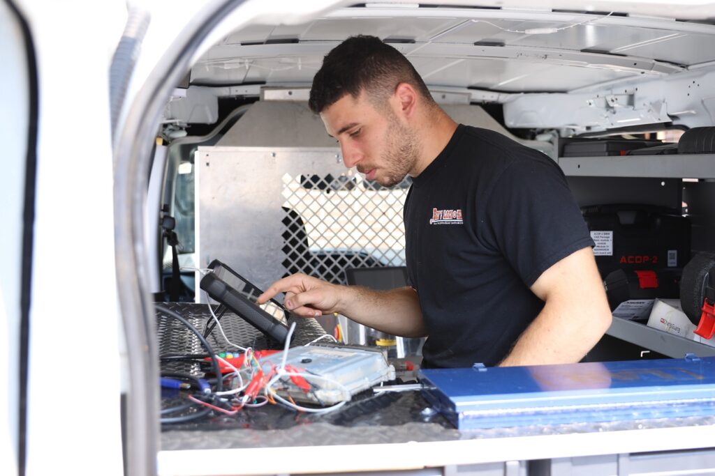 A local locksmith works inside a service van, using a tablet device near electronic equipment and wires on a workbench.