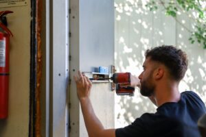 A local locksmith uses a power drill to install or repair a lock mechanism on a metal door. A fire extinguisher is mounted on the wall nearby.