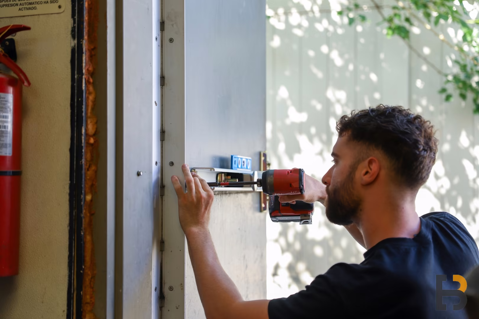 A local locksmith uses a power drill to install or repair a lock mechanism on a metal door. A fire extinguisher is mounted on the wall nearby.