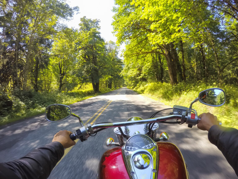 Motorcyclist riding a red motorcycle down a tree-lined road on a sunny day in New Jersey, with hands visible on the handlebars.