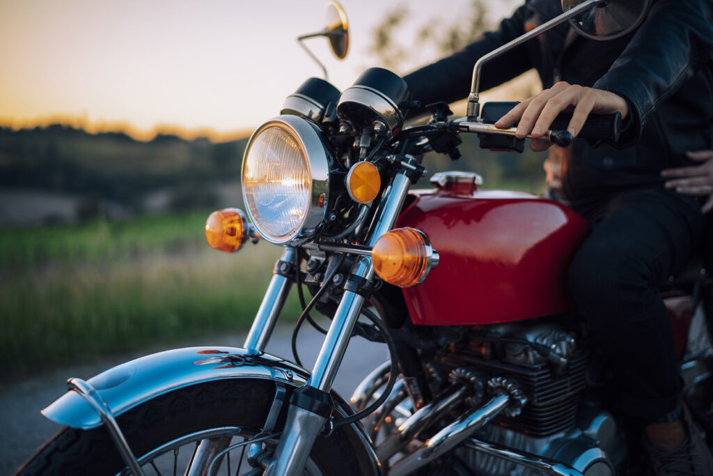 Close-up of a person riding a red motorcycle on a road with another person seated behind; background shows blurred greenery and a sunset sky, capturing the freedom of exploring local roads near me.